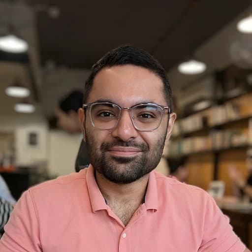 Hardeep at a café in a pink polo, holding coffee with a bookshelf behind